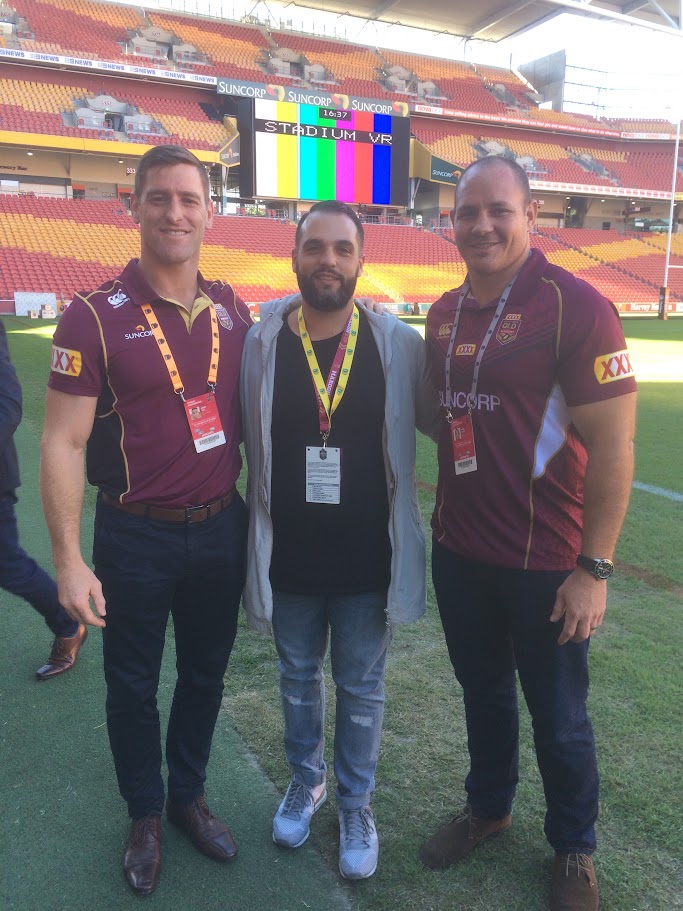 Two Queensland Maroons players in maroon jerseys standing on the field with a marketing team member before match day