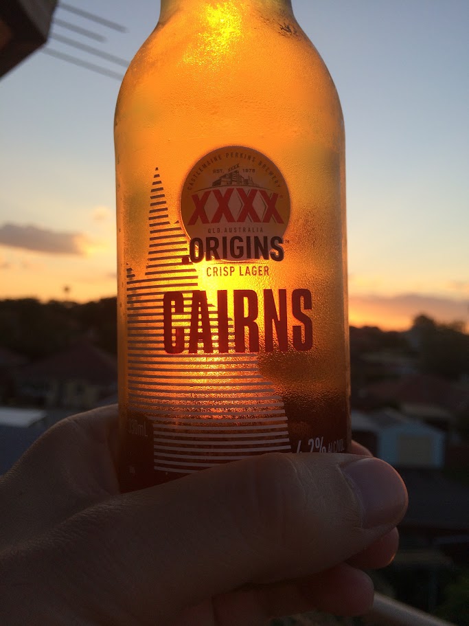 Close-up of a chilled XXXX Origins Crisp Lager bottle held up against a sunset sky